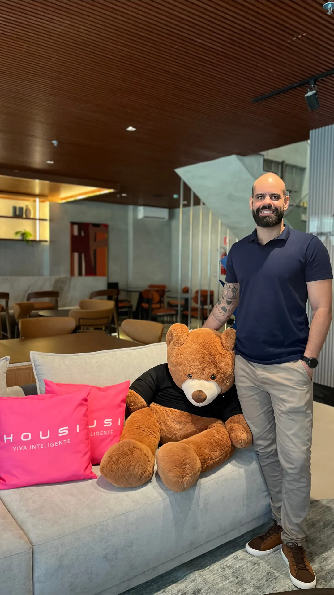 Fundador da ROTNA em pé no lobby de um empreendimento, ao lado de um urso de pelúcia gigante no sofá.