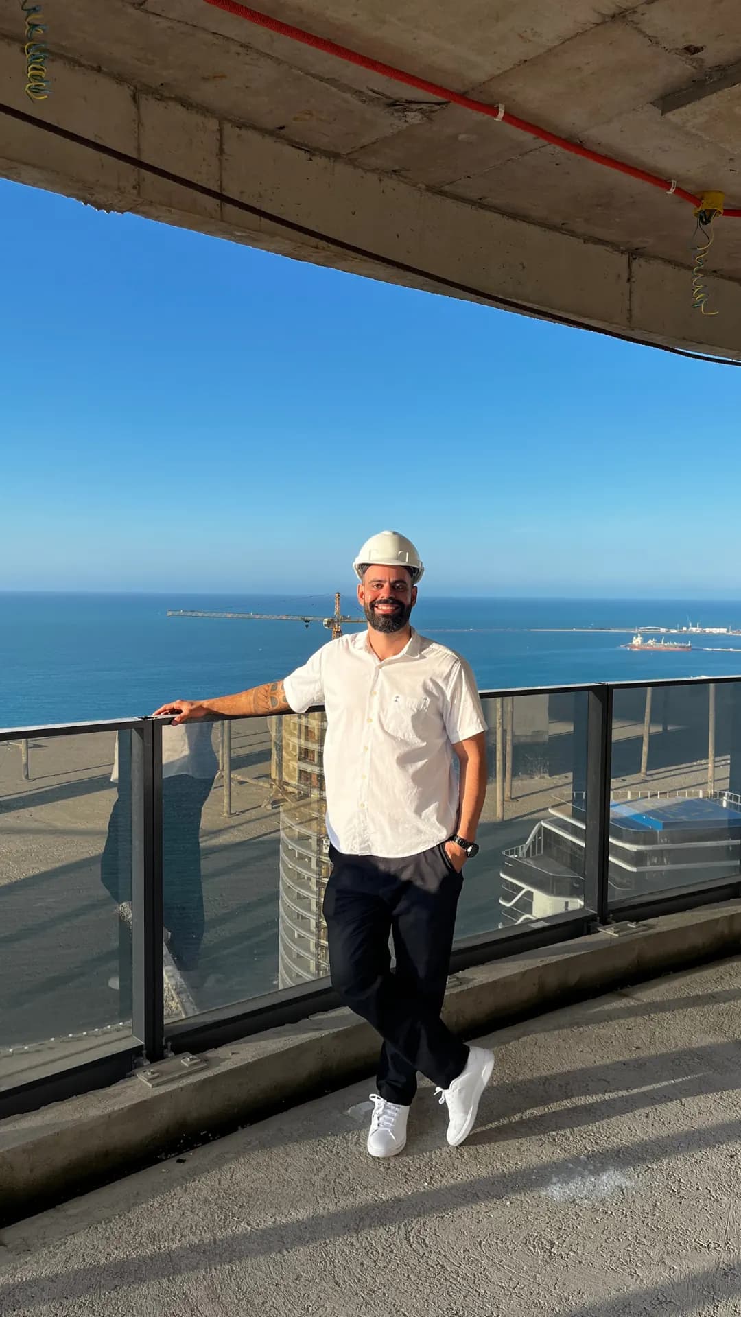 Fundador da ROTNA de capacete, apoiado em um guarda-corpo de vidro em uma obra, com o mar ao fundo.